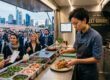 Köchin bereitet an einer offenen Streetfood-Theke ein Gericht zu, während Gäste vor der Frankfurter Skyline anstehen