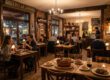 Gäste essen an Holztischen im warm beleuchteten Bistro „Le Jardin“ mit Kronleuchter