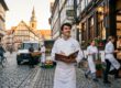 Koch mit Speisekarte steht vor einem Gasthof in einer Fachwerk-Altstadt, Kollegen tragen Speisen hinaus